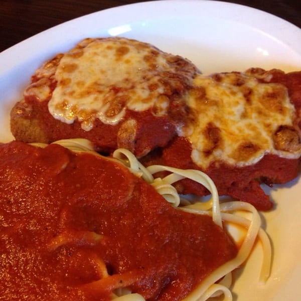 Chicken Parmesan with melted mozzarella cheese and red sauce, served alongside pasta on a white plate.
