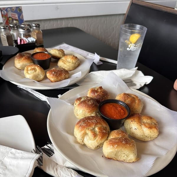 Two plates of La Famiglia's garlic knots with marinara sauce, salt and pepper shakers, and a drink.