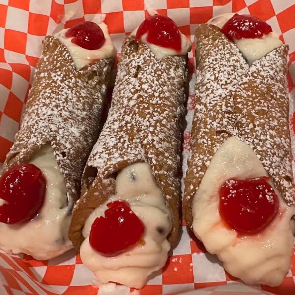 Three cannolis with powdered sugar, white cream filling, and a cherry on red and white checkered paper.